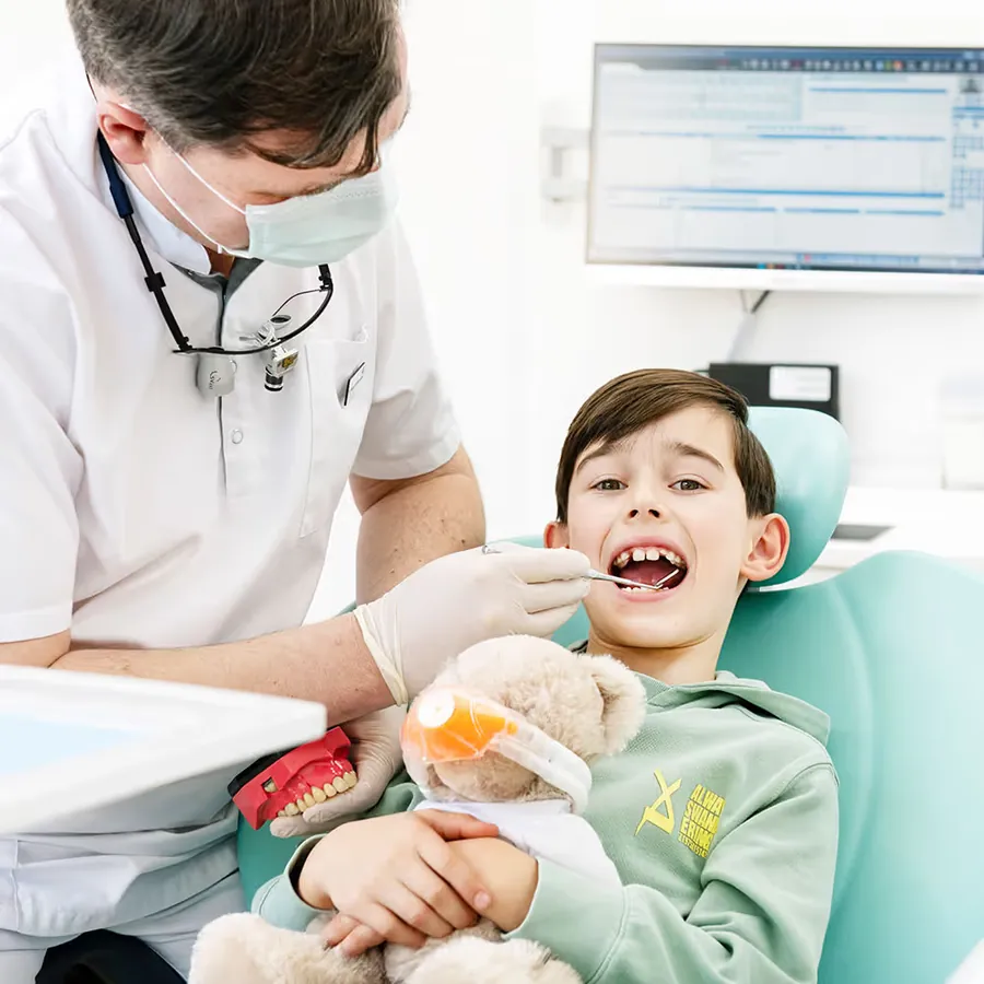 Zahnarzt untersucht einen lachenden Jungen, der einen Teddybären im Arm hält